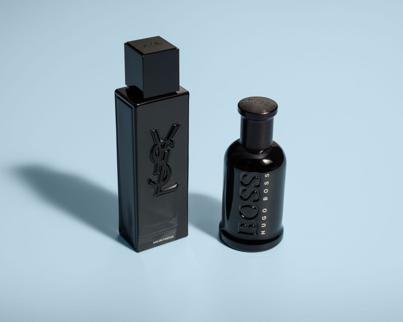 Voici deux parfums élégants sur un fond bleu clair : un flacon noir rectangulaire de Yves Saint Laurent et un flacon noir arrondi de Hugo Boss.