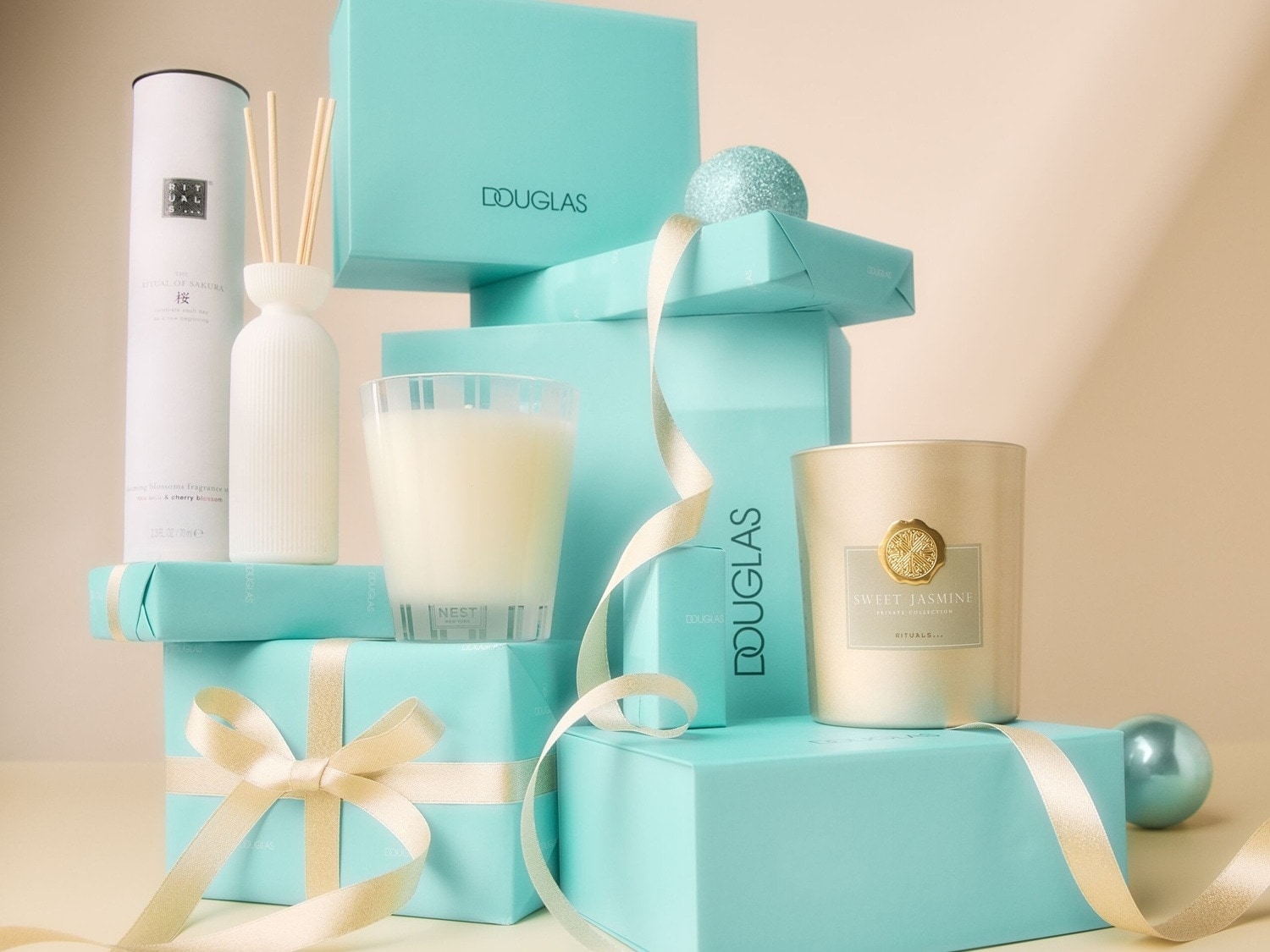 Scatole regalo turchesi Douglas impilate con candele profumate e diffusore di fragranze Rituals, avvolte da un nastro dorato.