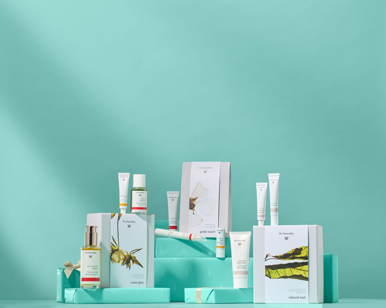 Scopri la collezione Dr. Hauschka con confezioni regalo 'warm glow' e 'relaxed soul', creme e oli essenziali presentati su un elegante sfondo turchese.