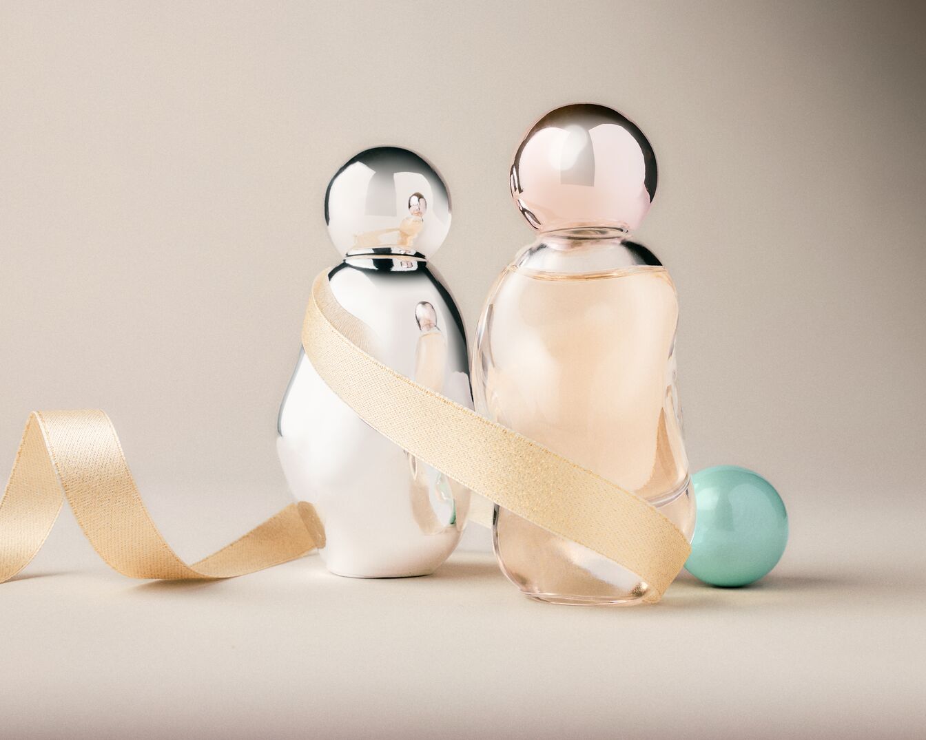 Due flaconi di profumo, uno argentato e l'altro trasparente con tappo rosa, avvolti da un nastro dorato su sfondo neutro.