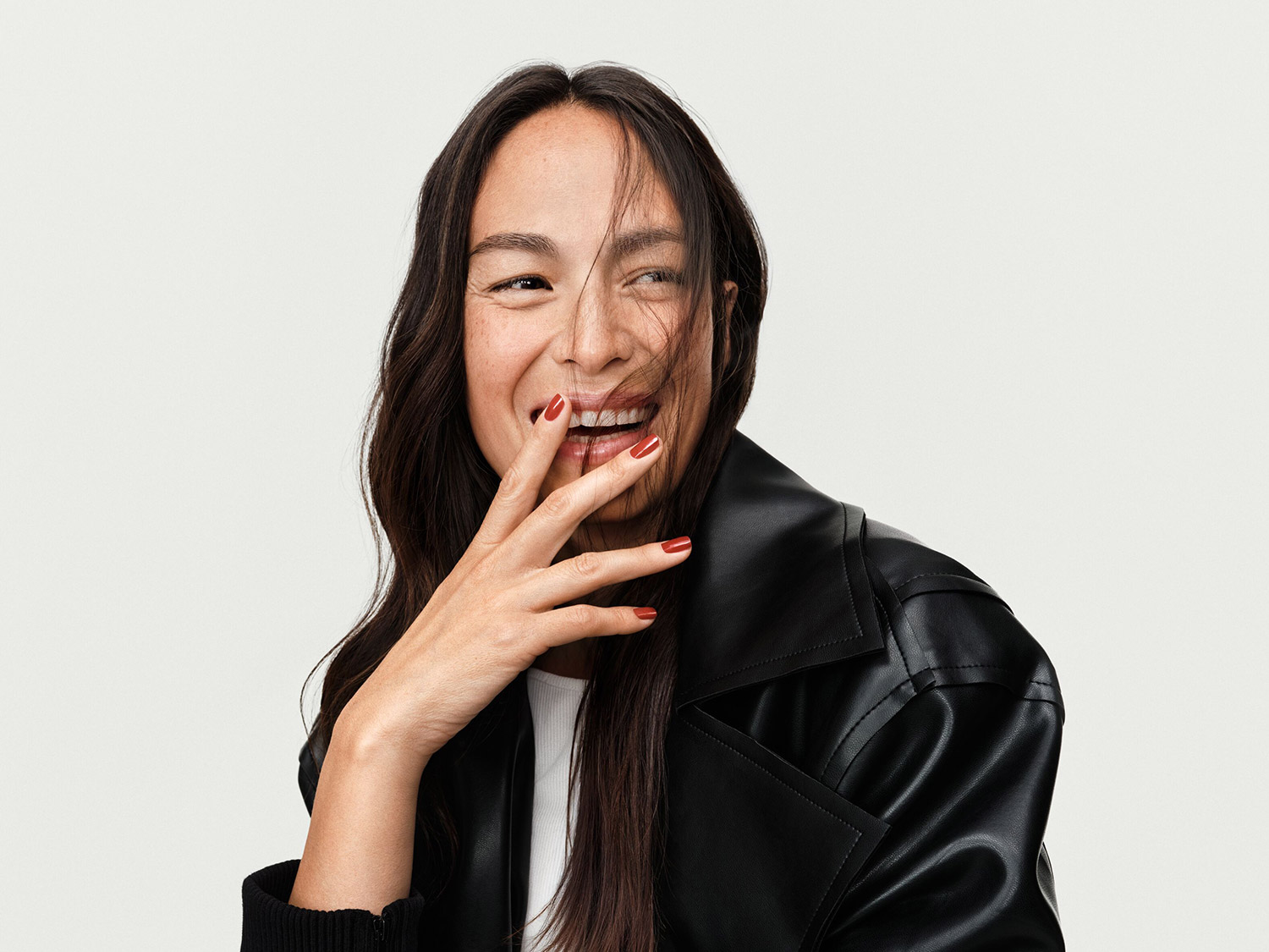 Une femme souriante, les doigts ornés de vernis rouge vif, porte une veste en cuir noire élégante, un look à la fois moderne et audacieux.