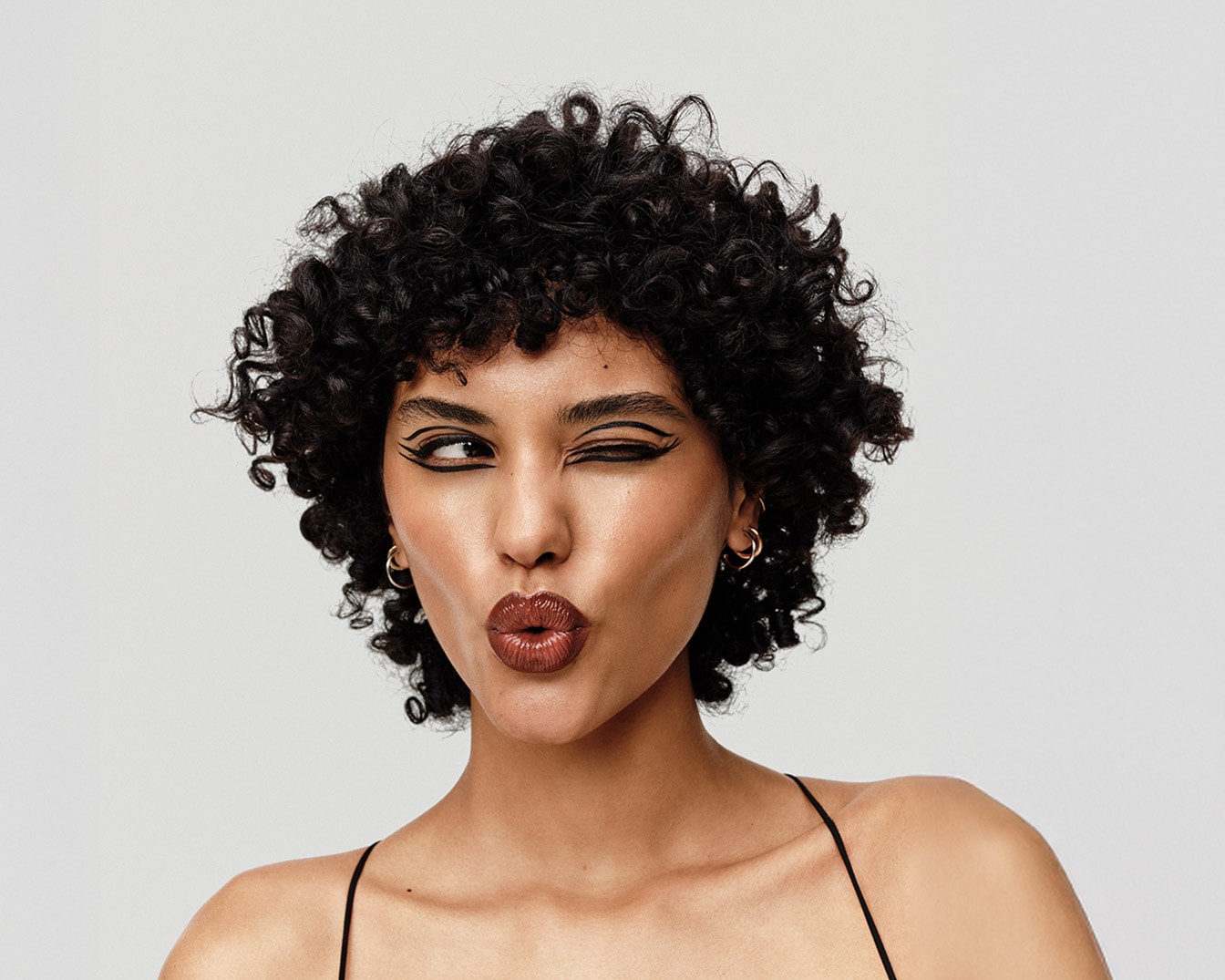 Une femme aux cheveux bouclés et au maquillage audacieux, avec un eye-liner graphique noir et des lèvres brillantes, fait un clin d'œil espiègle.