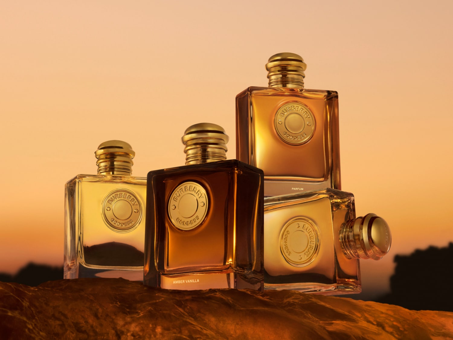 Cuatro frascos de perfume Burberry Goddess, dos de color ámbar claro y dos de color ámbar oscuro, con tapas doradas y un sello dorado en el centro, se exhiben sobre una superficie texturizada de color marrón rojizo, con un cielo naranja y amarillo de fondo.