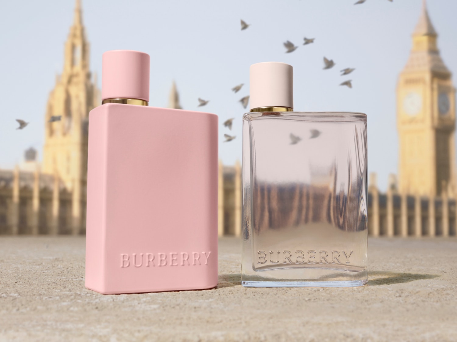 Dos frascos de perfume Burberry, uno rosa opaco y otro transparente, se alzan sobre un fondo borroso de edificios históricos y pájaros volando, evocando la esencia de Londres.