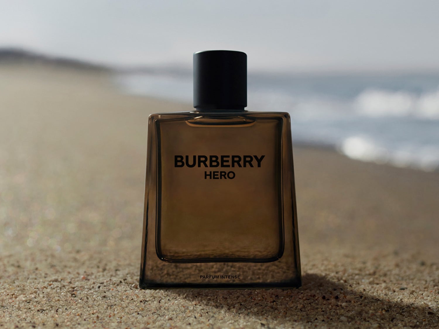 El perfume Burberry Hero Parfum Intense, en un frasco de vidrio ahumado con tapa negra, se alza sobre la arena de una playa, con el mar de fondo.