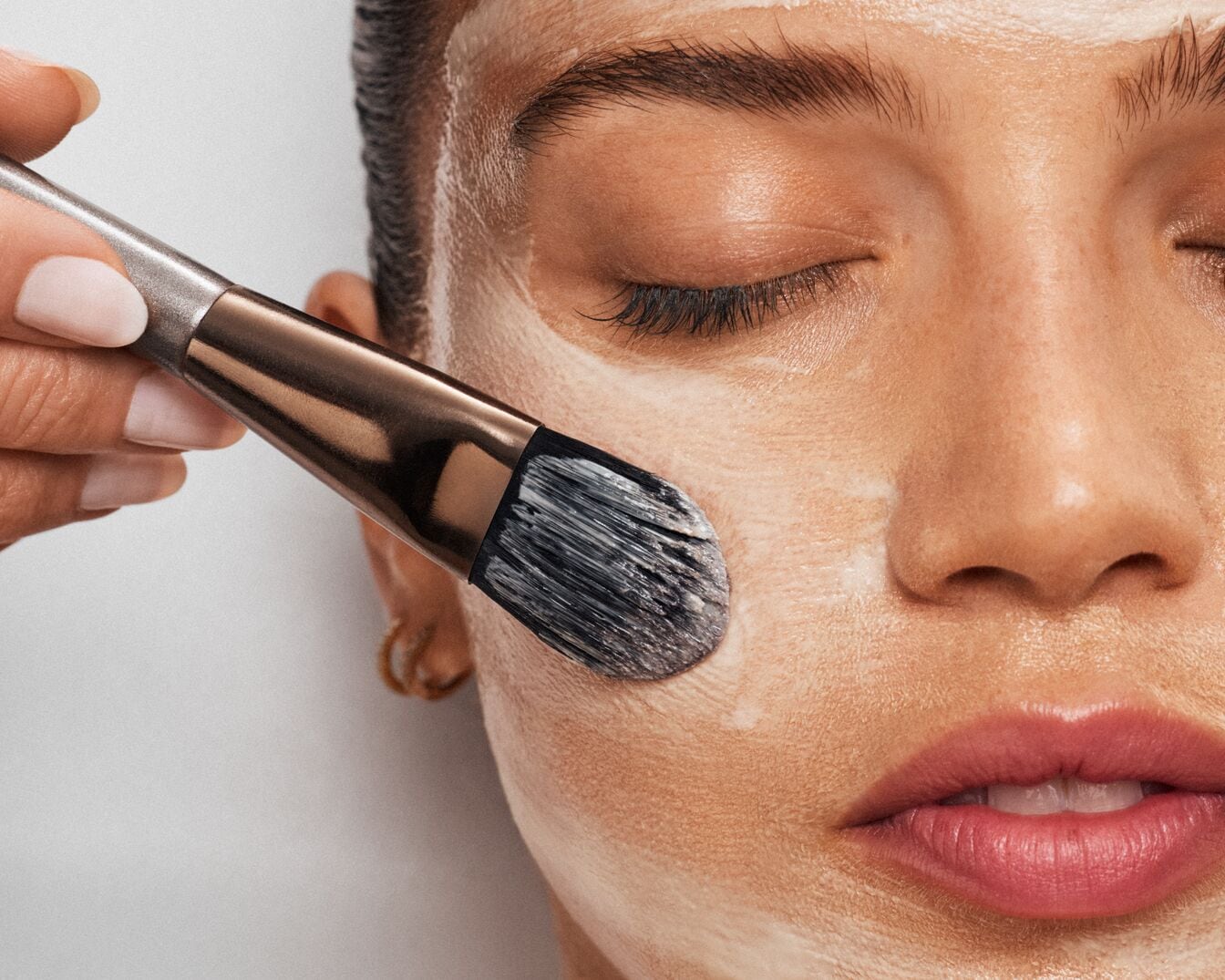 Nahaufnahme einer Frau, die mit einem Pinsel eine weiße Gesichtsmaske aufträgt, um ihre Haut zu pflegen und zu verjüngen.