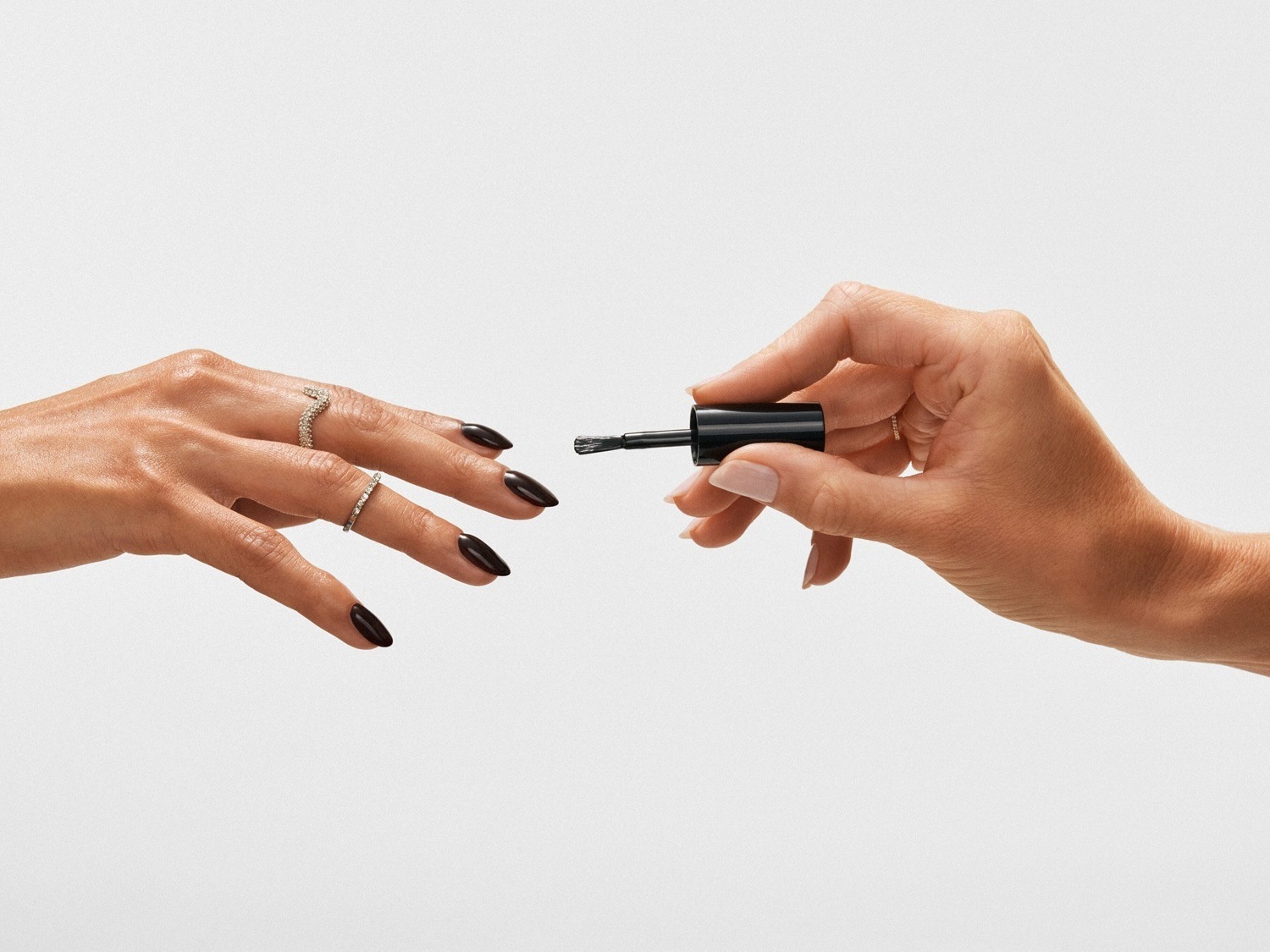 Dłoń z ciemnym manicure i pierścionkami zbliża się do dłoni trzymającej pędzelek z czarnym lakierem do paznokci, gotowa na aplikację.
