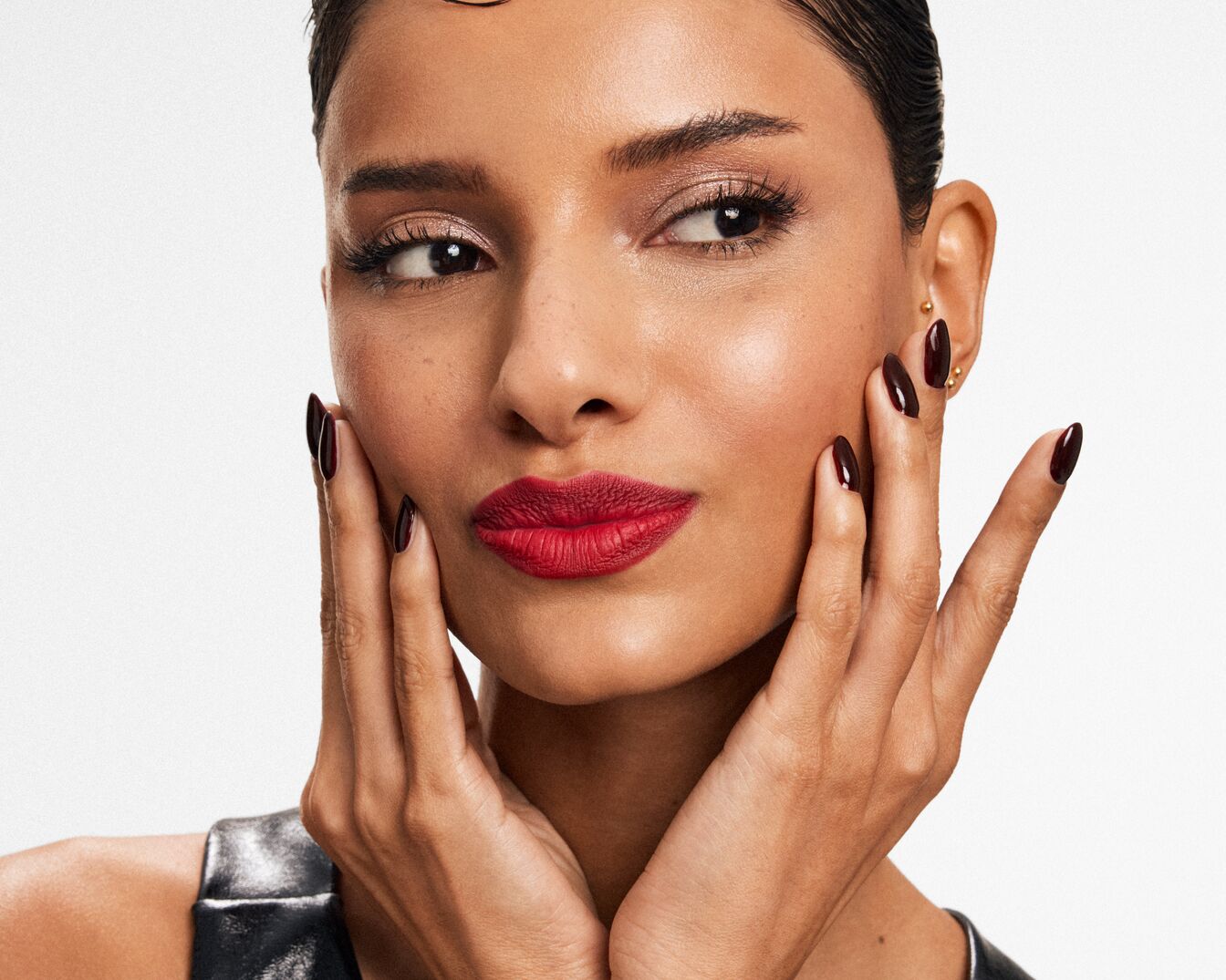 Nahaufnahme einer Frau mit roten Lippen, glänzenden Lidschatten und dunkelroten Nägeln, die ihr Gesicht berührt und einen eleganten Look präsentiert.