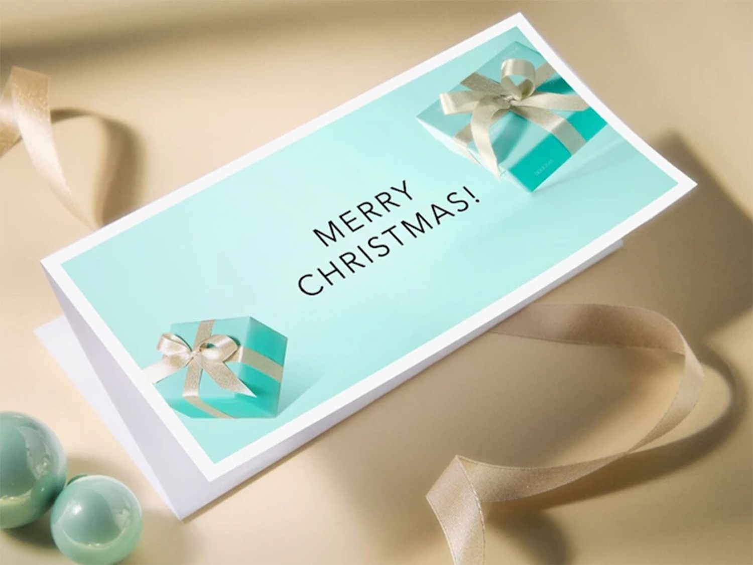 Biglietto d'auguri con la scritta 'Buon Natale!' in nero, decorato con due pacchetti regalo turchesi con fiocchi color crema e palline decorative turchesi.