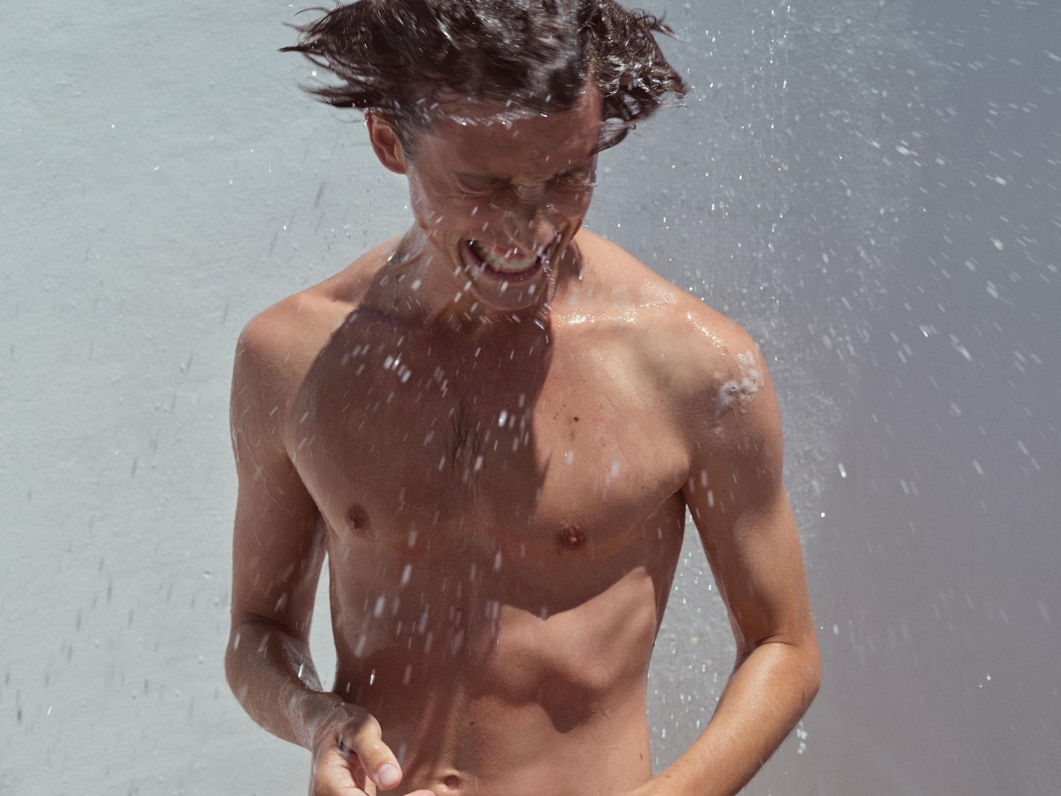 Ein junger Mann mit dunklen, nassen Haaren und freiem Oberkörper steht unter einer Dusche und genießt die Erfrischung.