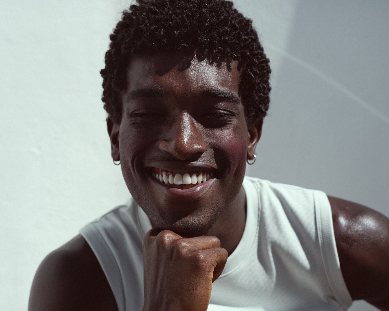 Ein lächelnder, dunkelhäutiger Mann mit krausem Haar und einem weißen Tanktop posiert vor einer weißen Außenwand.