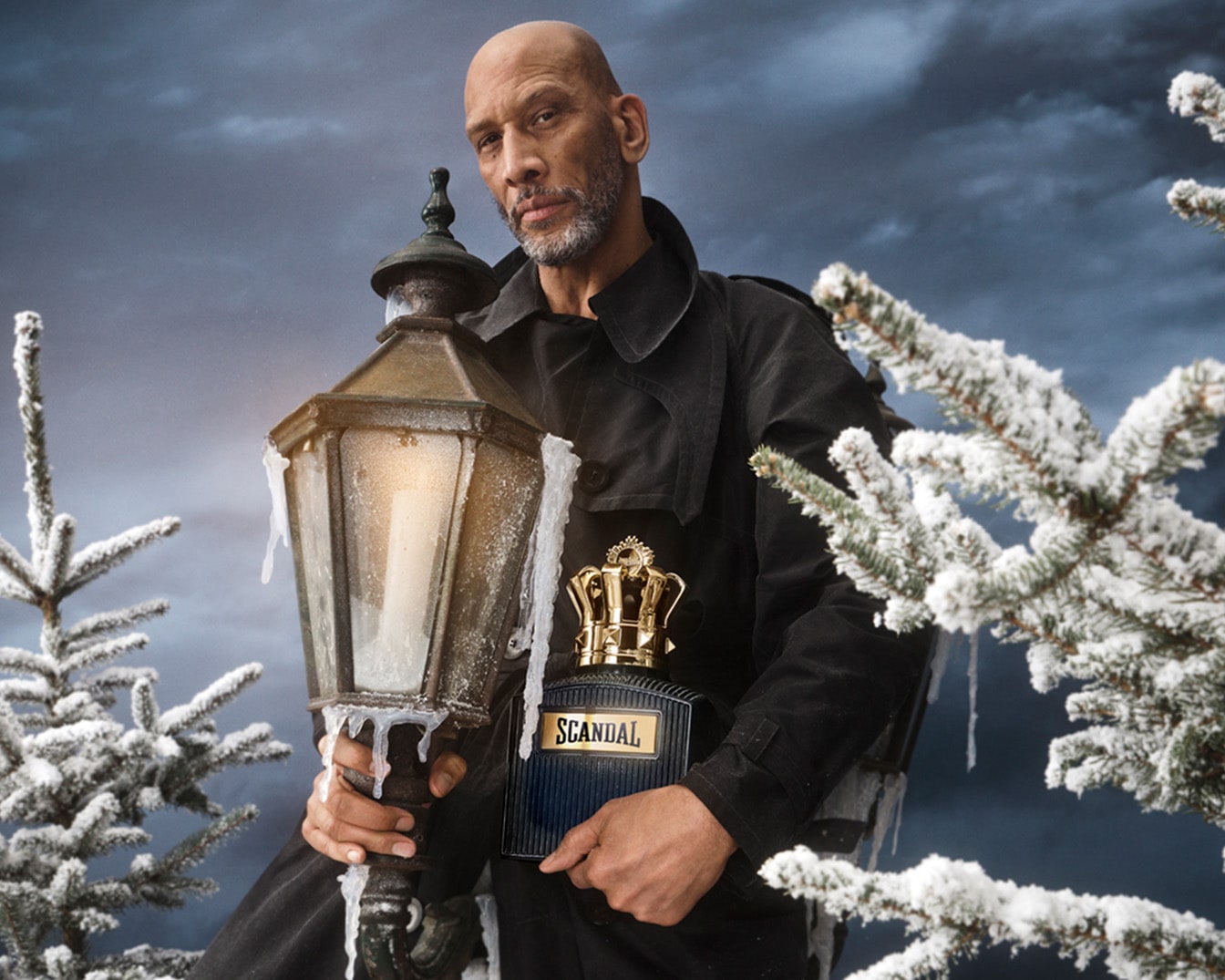 Vyras juodu paltu laiko žibintą ir „Scandal“ kvepalus su auksiniu karūnos dangteliu apsnigtame fone.