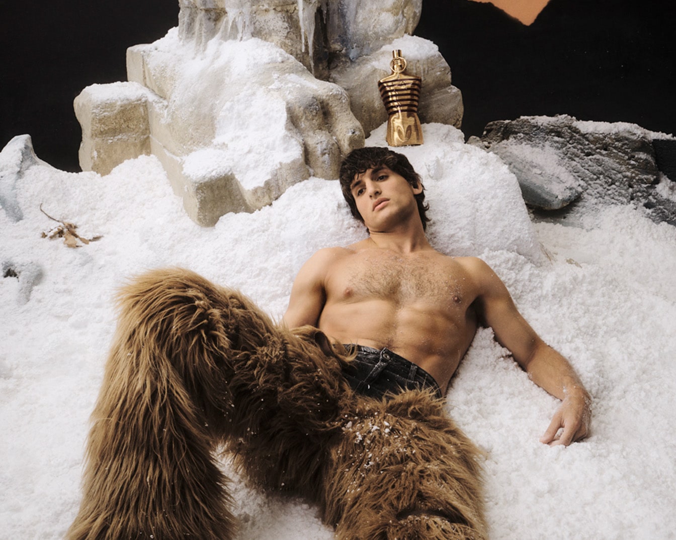 Ant sniego guli vyras su kailinėmis kelnėmis, o šalia jo stovi auksinis kvepalų buteliukas.