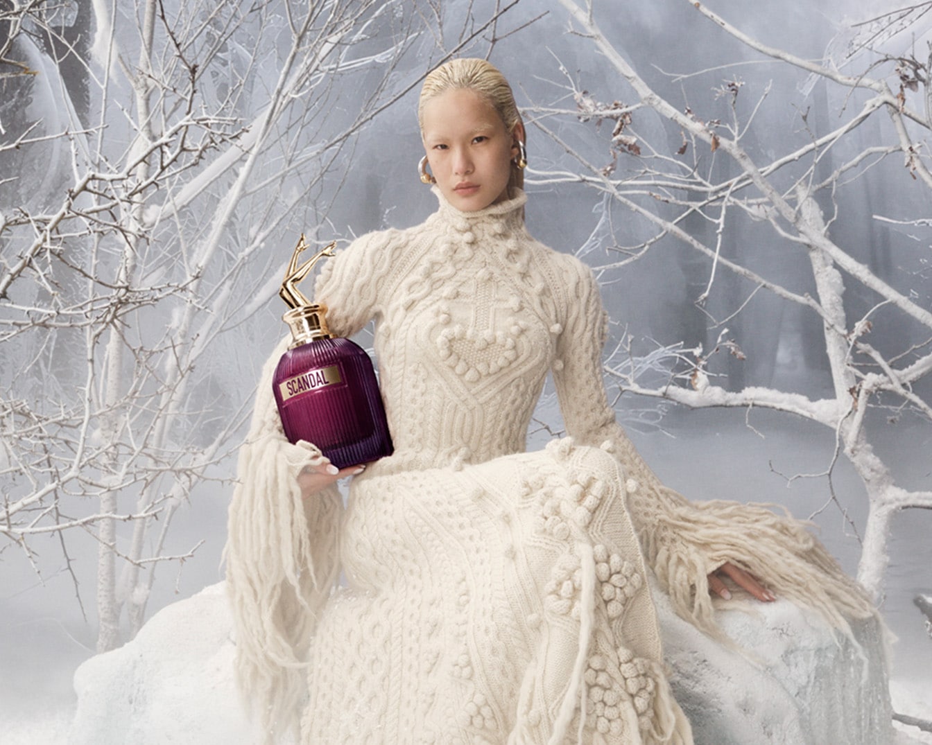 Sniego fone moteris su baltu megztiniu laiko burgundišką „Scandal“ kvepalų buteliuką su aukso spalvos dangteliu.