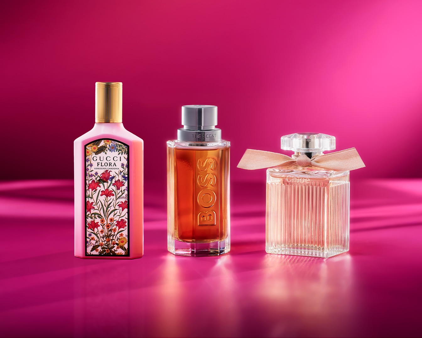 Eine Kollektion von Damendüften, darunter Gucci Flora mit rosa Flakon und Blumenmuster, Boss The Scent mit bernsteinfarbenem Flakon und Chloé mit geripptem Flakon und Schleife, alle vor einem leuchtend rosa Hintergrund.