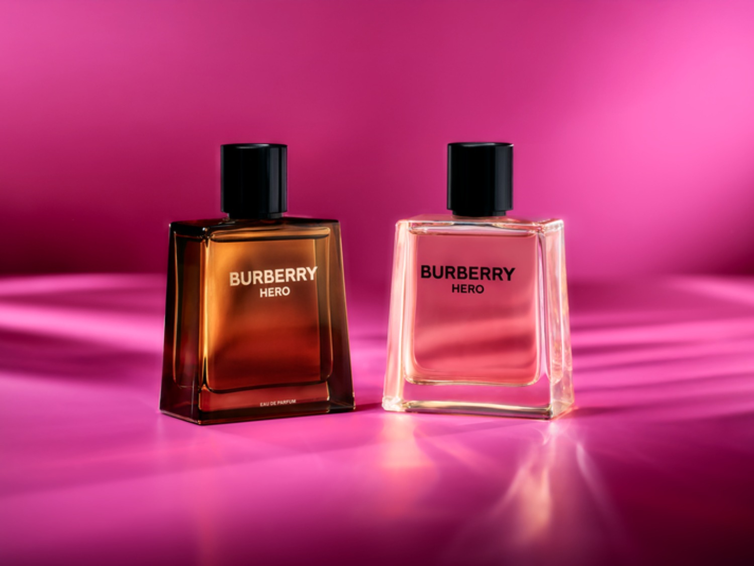Zwei Burberry Hero Parfümflaschen stehen vor einem pinkfarbenen Hintergrund, eine mit bernsteinfarbenem und die andere mit rosa Duft.