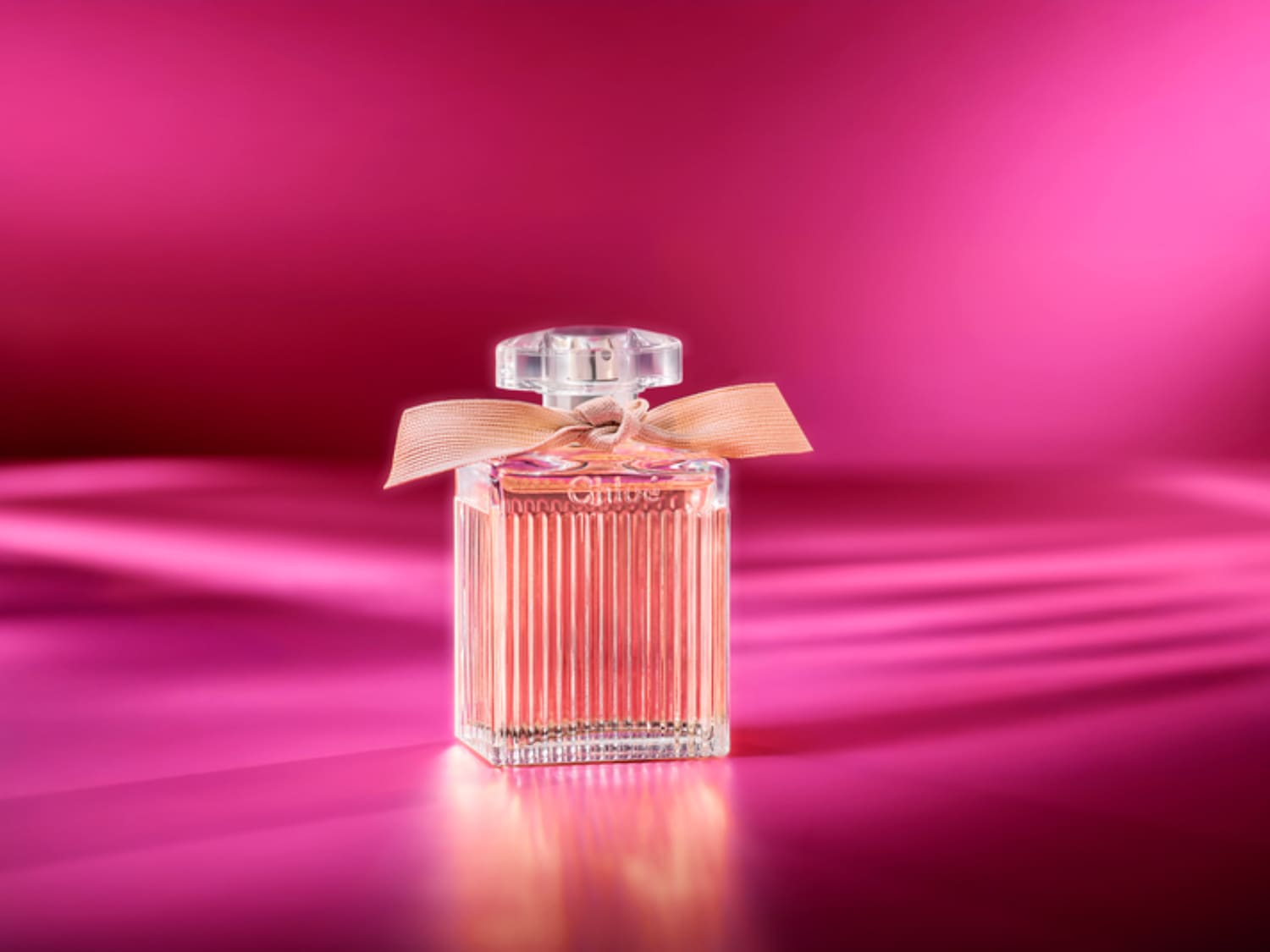 Ein rechteckiger, durchsichtiger Glasflakon mit rosa Flüssigkeit, verziert mit einer zarten Schleife, steht vor einem leuchtend pinkfarbenen Hintergrund.