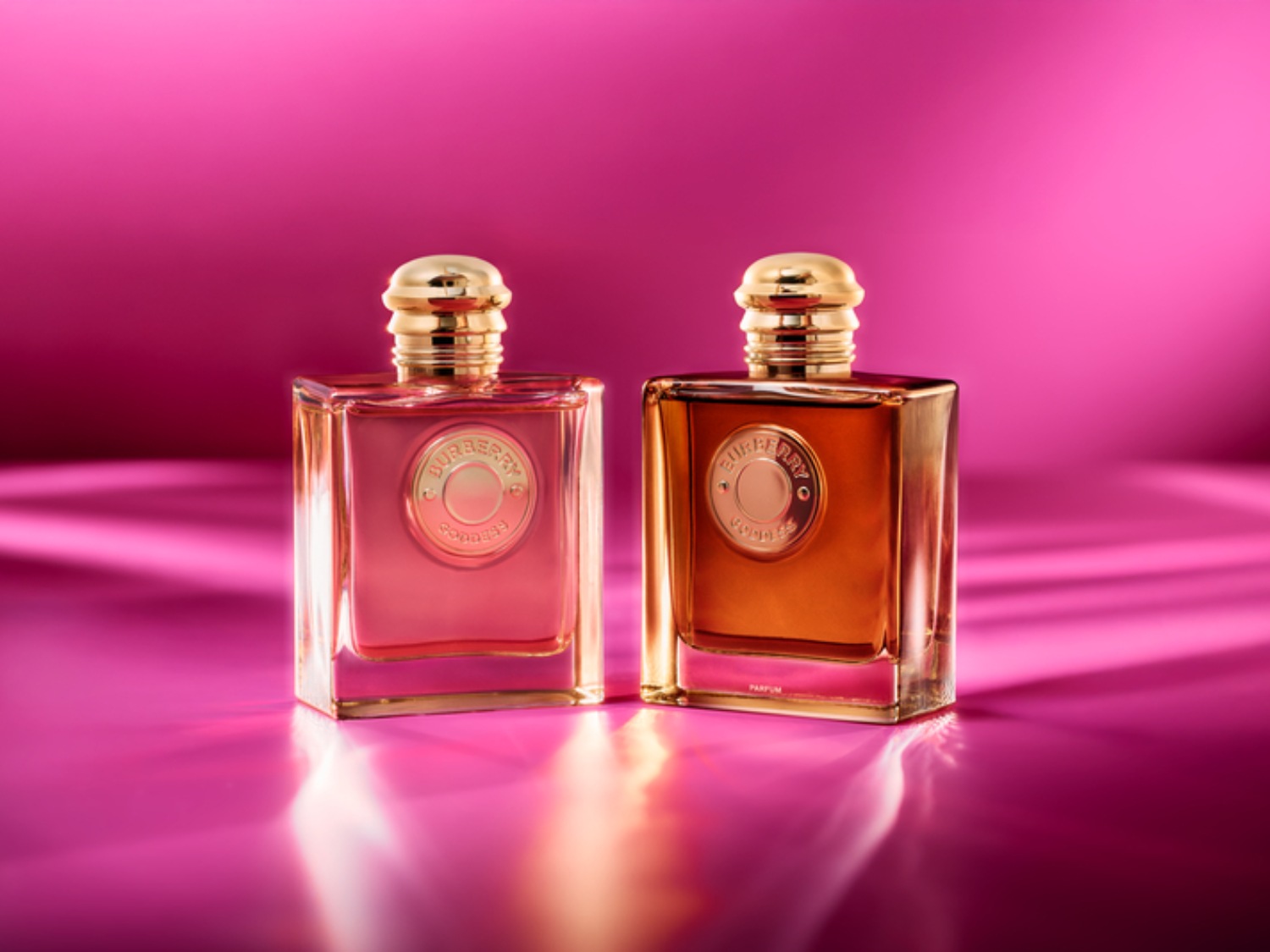 Zwei Burberry Goddess Parfümflaschen stehen vor einem pinkfarbenen Hintergrund, die eine mit einem rosa Farbton und die andere mit einem bernsteinfarbenen Farbton.