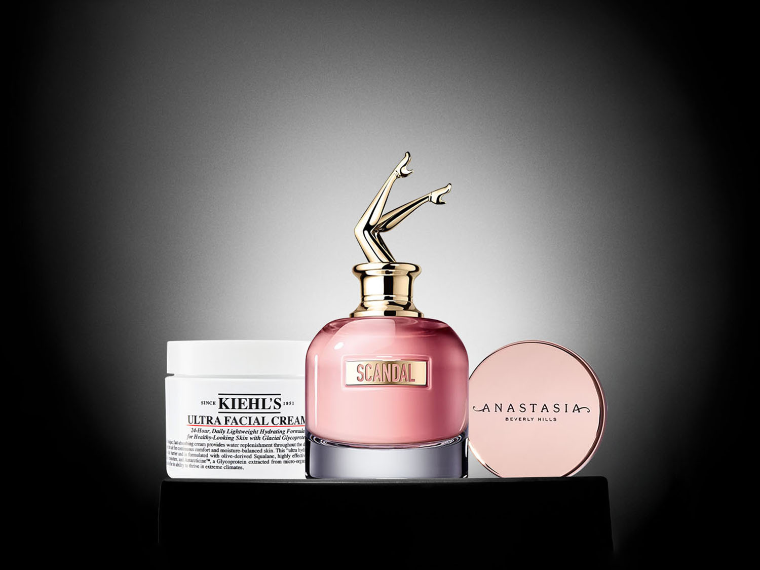 Op een zwarte achtergrond staan drie schoonheidsproducten: Kiehl's Ultra Facial Cream, Scandal parfum met een gouden dop in de vorm van benen, en Anastasia Beverly Hills compact poeder.