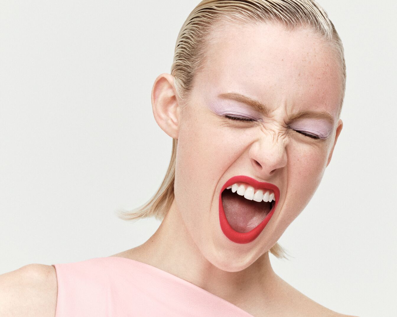 Egy nő, rövid szőke hajjal, rózsaszín felsőben, rózsaszín szemhéjfestékkel és élénkpiros rúzzsal, csukott szemmel és tátott szájjal látható.