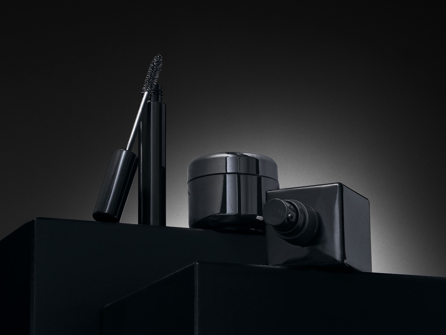 Juodos spalvos kosmetikos rinkinys, įskaitant tušą, kremo indelį ir buteliuką ant juodo paviršiaus.