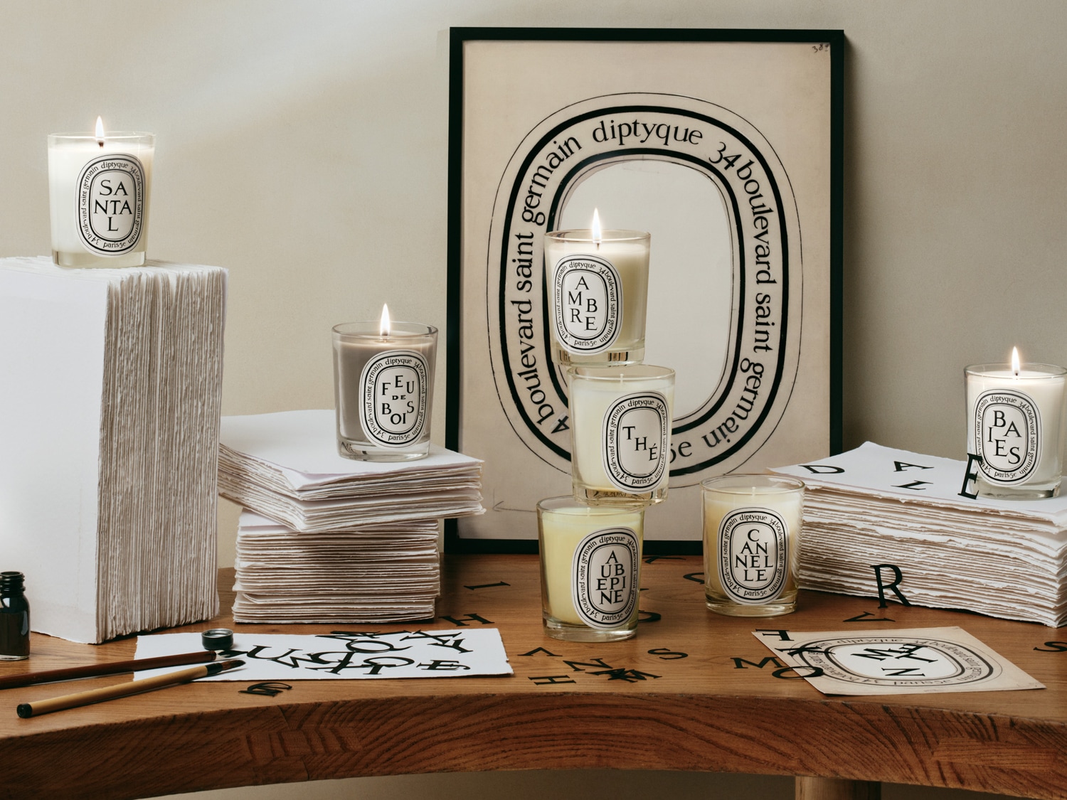Eine Sammlung von Diptyque-Duftkerzen mit verschiedenen Düften wie Santal und Ambre, die auf einem Holztisch mit Papierstapeln und Schreibutensilien arrangiert sind.