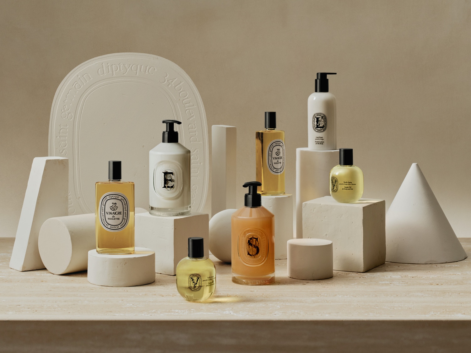 Eine Sammlung von Diptyque-Produkten, darunter Bodyspray, Bodylotion und Handseife, präsentiert auf Podesten in verschiedenen Formen auf strukturiertem Untergrund.