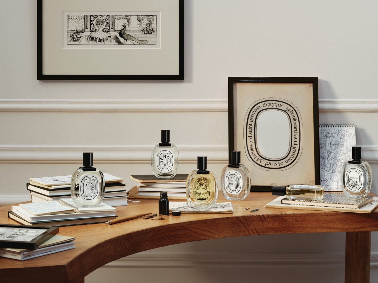 Mehrere Diptyque-Parfumflaschen mit schwarzen Verschlüssen sind auf einem Holztisch zwischen Notizbüchern und Stiften angeordnet, mit einem gerahmten Bild und einer Illustration im Hintergrund.
