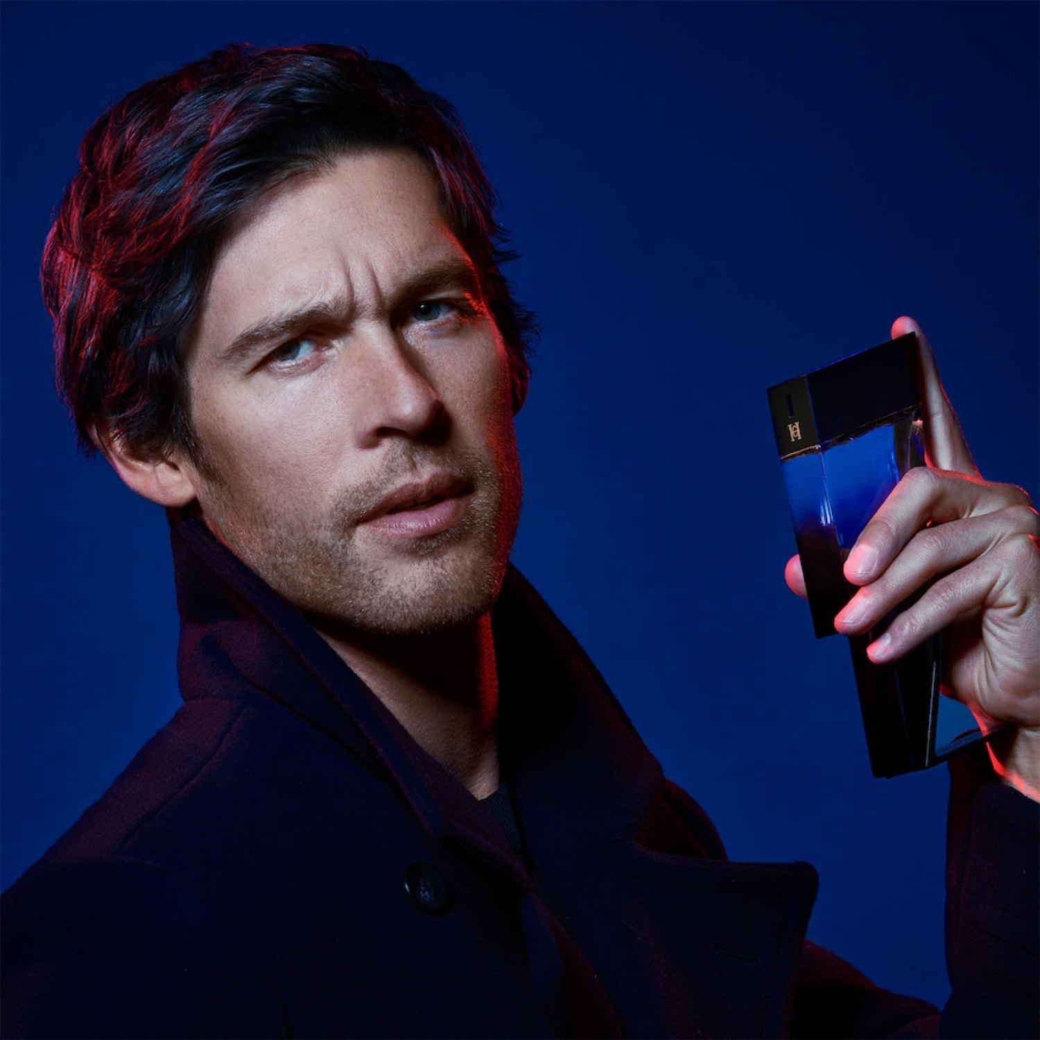 Na ciemnoniebieskim tle mężczyzna w ciemnym płaszczu trzyma flakon perfum Carolina Herrera o gradientowym, niebiesko-czarnym kolorze.