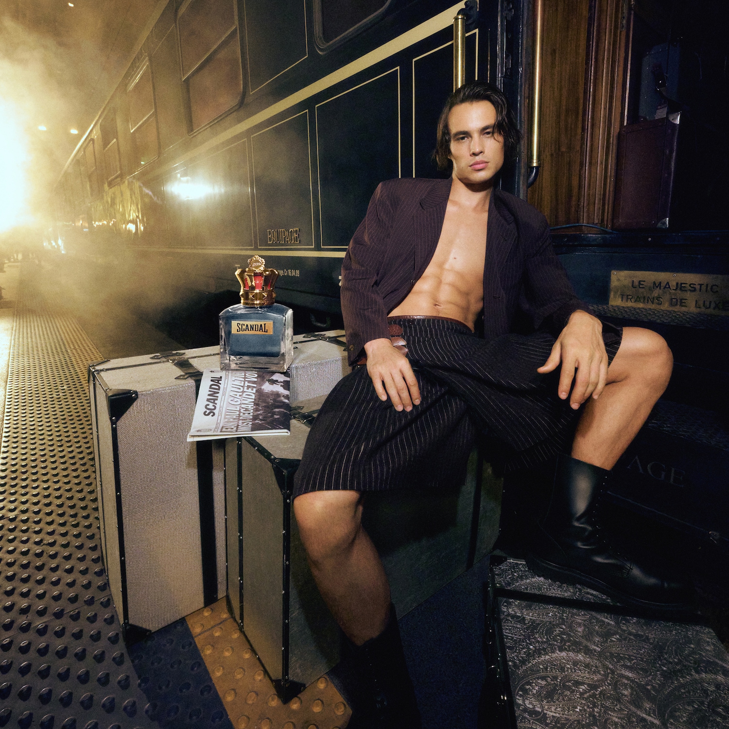 Na peronie, mężczyzna w rozpiętej marynarce siedzi na walizkach obok flakonu perfum Scandal, tworząc odważny i luksusowy wizerunek.