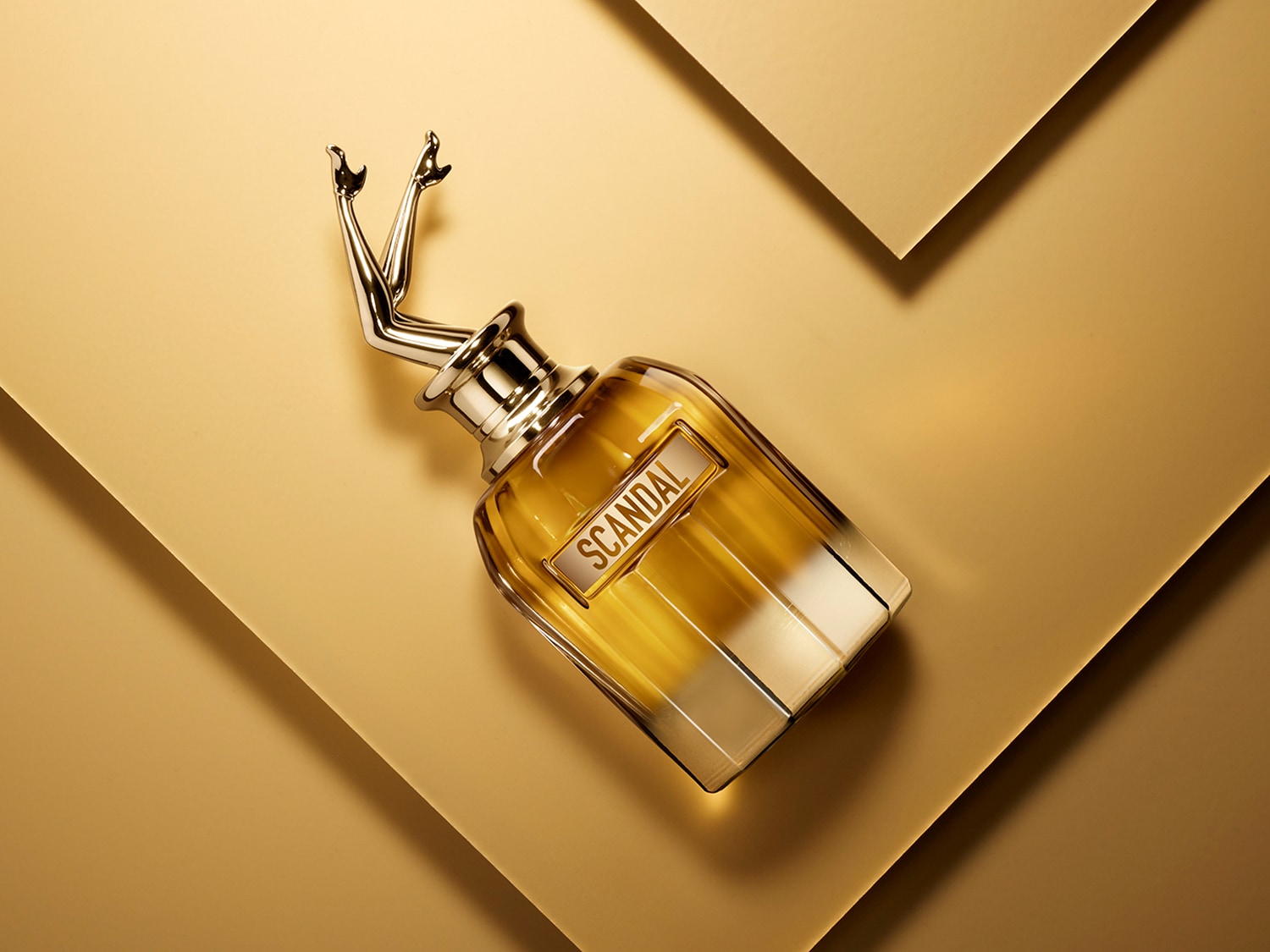 Butelka perfum Scandal z przezroczystego szkła z złotymi akcentami i złotymi nogami skierowanymi do góry, umieszczona na złotym tle.