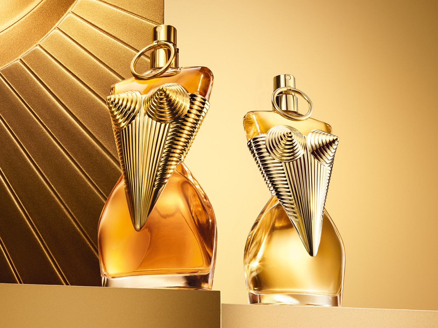 Dwa flakony perfum Jean Paul Gaultier w kształcie kobiecego torsu, jeden z bursztynowym płynem, drugi z jaśniejszym, oba z metalicznymi, złotymi detalami na złotym tle.