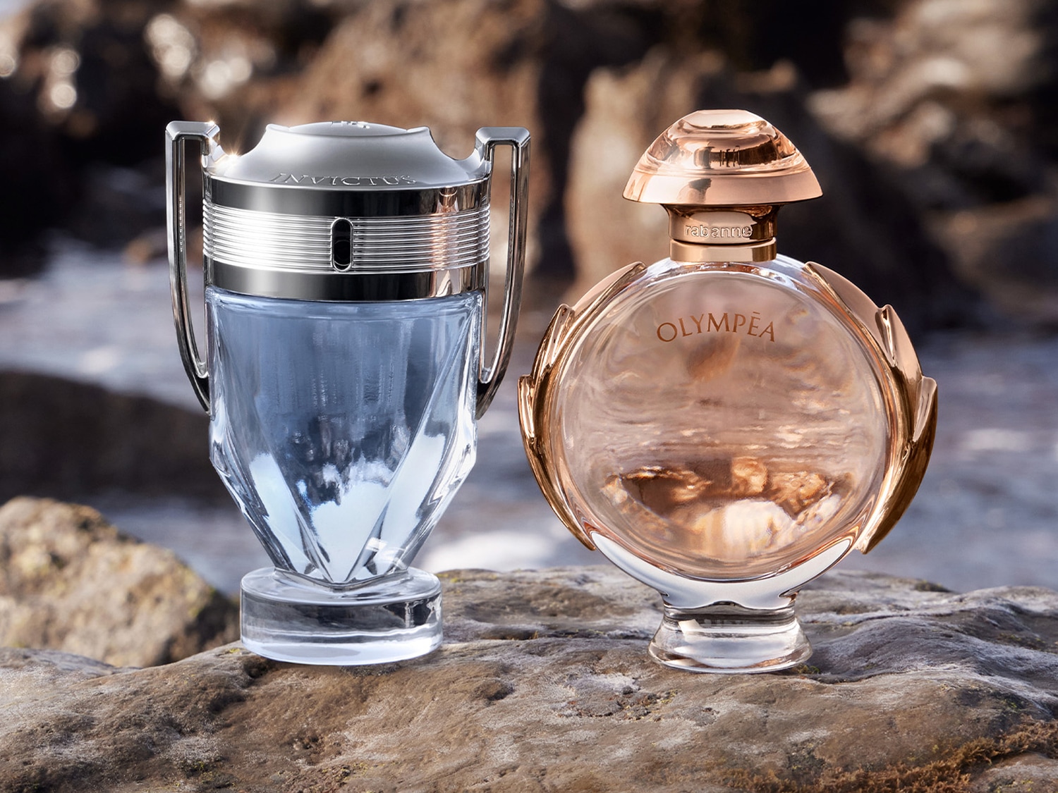 Dwa flakony perfum: Invictus, w kształcie pucharu, z niebieską cieczą, oraz Olympéa, okrągły, z różowo-złotymi akcentami, stoją na kamiennej powierzchni.