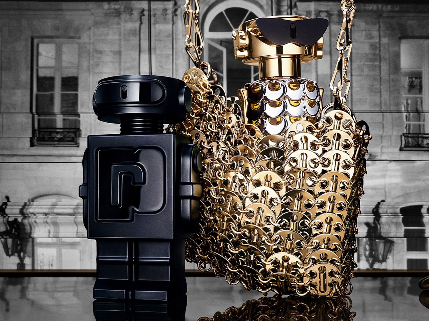Dwa flakony perfum w kształcie robotów: jeden czarny, matowy, drugi złoty, błyszczący, z metalową siatką i łańcuszkiem na tle budynku.