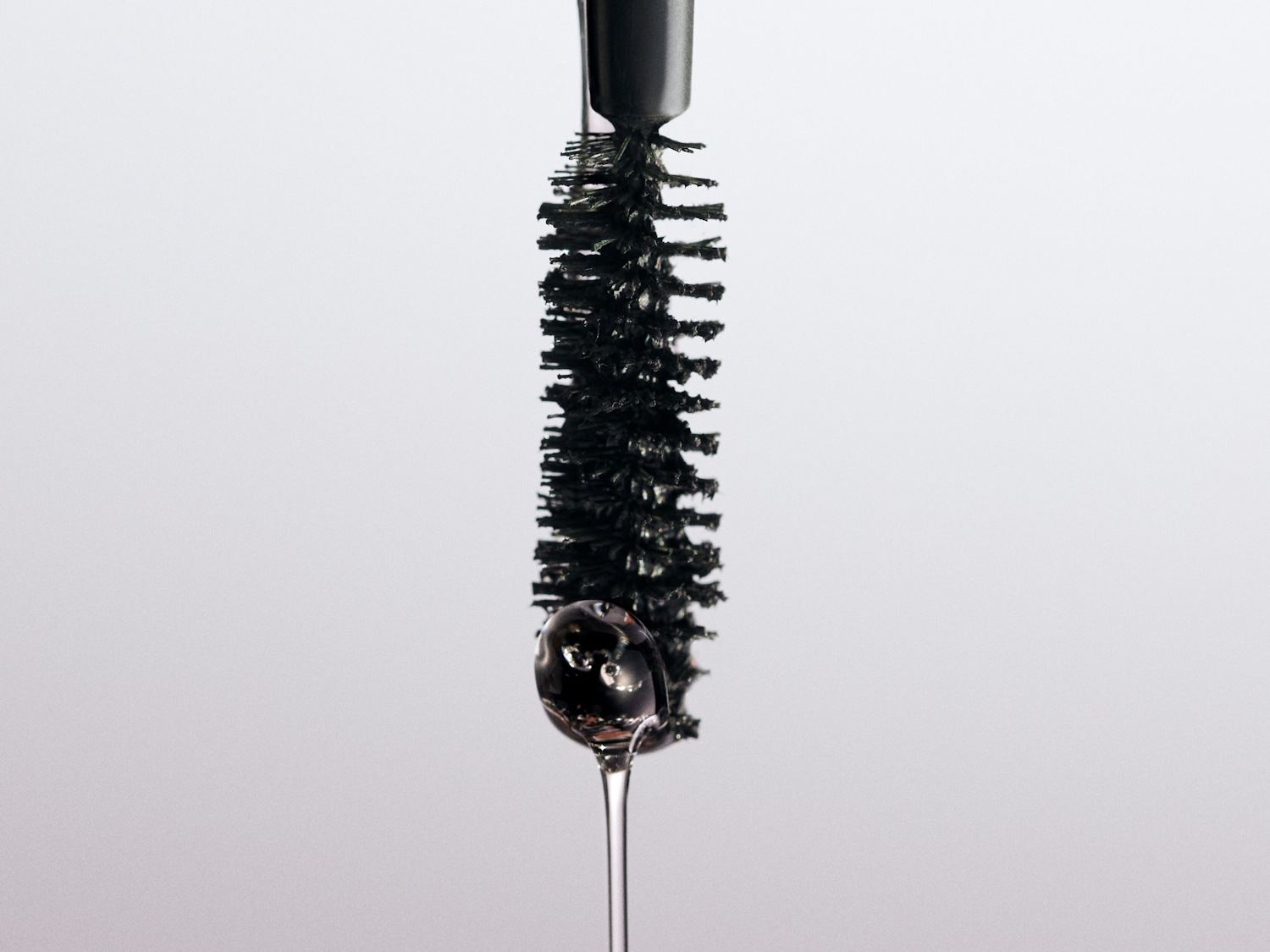 Close-up van een zwarte mascara borstel met een druppel heldere vloeistof die eraf valt, tegen een lichte achtergrond.