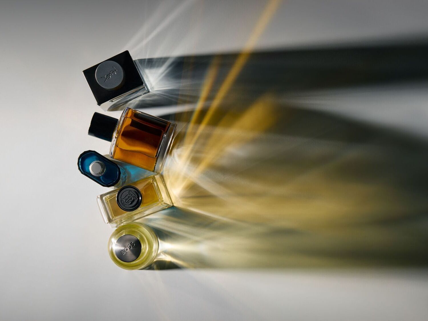 Įvairių kvepalų buteliukų kolekcija suprojektuota ant balto paviršiaus, apšviesta ryškiais šviesos spinduliais, sukuriančiais dramatiškus šešėlius.