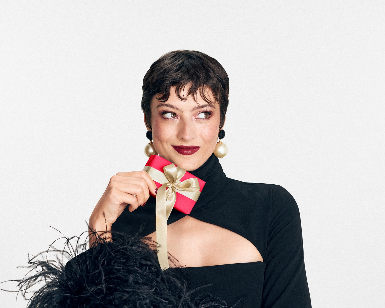 Une femme élégante en robe noire tient une carte-cadeau rouge ornée d'un nœud doré, parfaite pour offrir un moment de beauté.