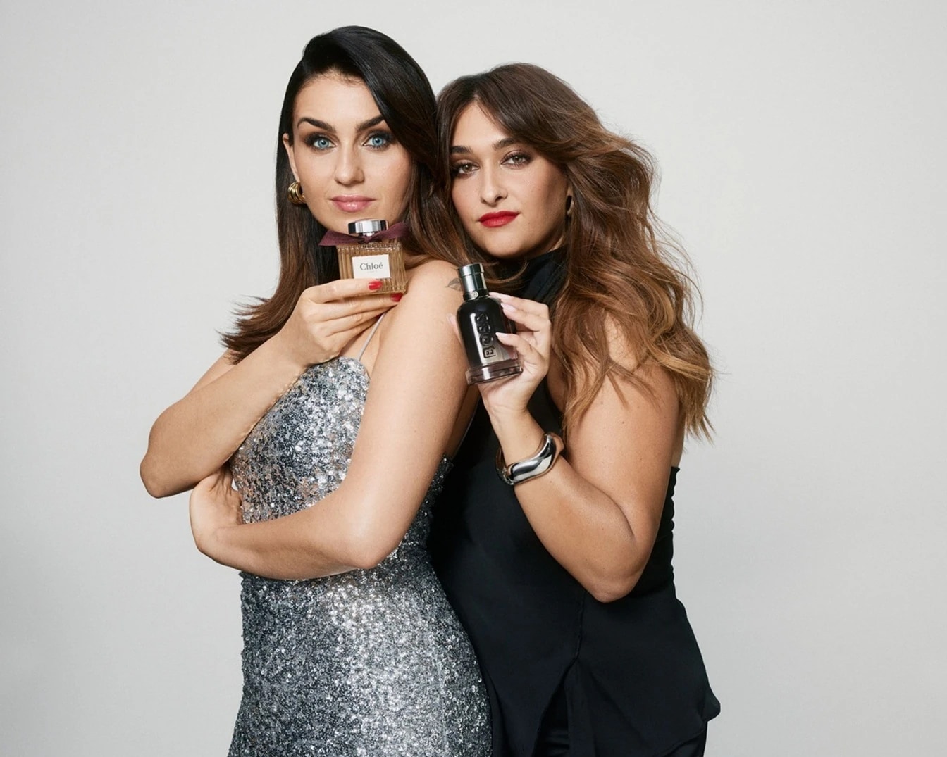 Zwei Frauen posieren mit Parfümflaschen: Die eine hält Chloé mit einer Schleife, die andere Boss in einer dunklen Flasche.