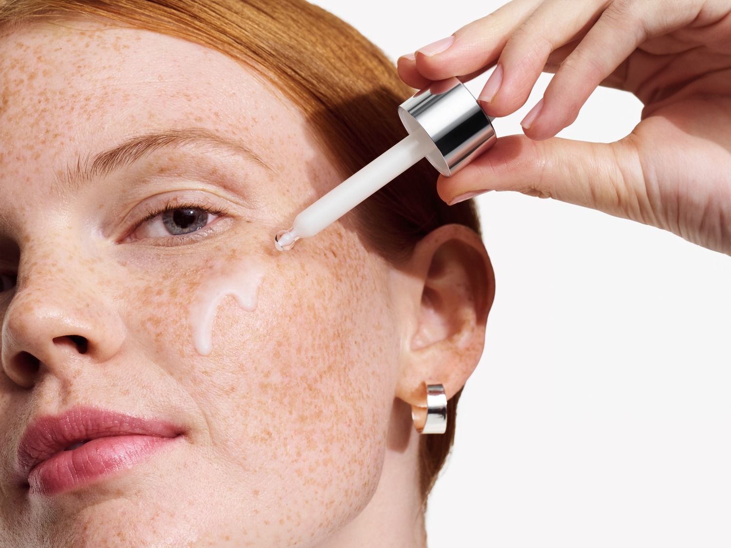 Een vrouw met rood haar en sproeten brengt een helder serum aan op haar gezicht met een witte applicator.