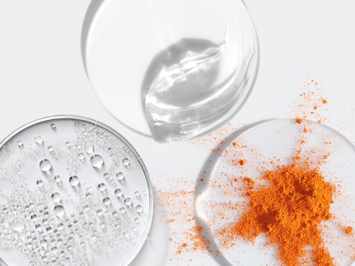 Gros plan sur trois coupelles transparentes contenant un gel clair, des gouttelettes d'eau et une poudre orange, offrant un aperçu des textures et des ingrédients.