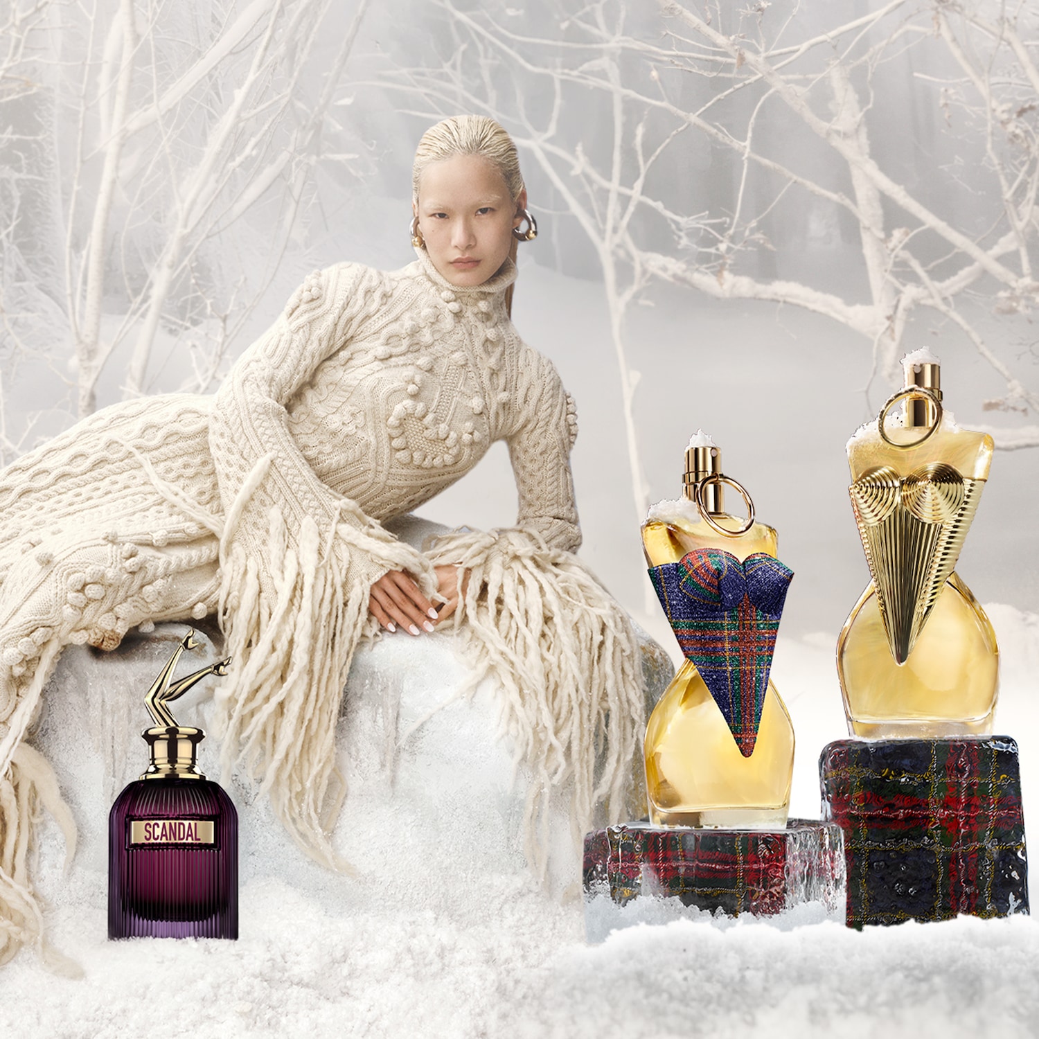 Découvrez une collection de parfums dans un décor hivernal, avec des flacons aux formes uniques et des détails dorés, présentés aux côtés d'un mannequin en tenue de maille.