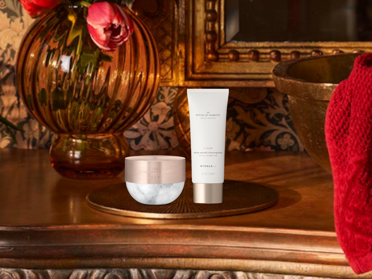 „Ritual of Namaste Day“ kremas marmuriniame indelyje su rožinio aukso spalvos dangteliu ir „Rituals...“ valomasis veido putos baltame buteliuke su rožinio aukso spalvos dangteliu ant rudos medinės komodos.