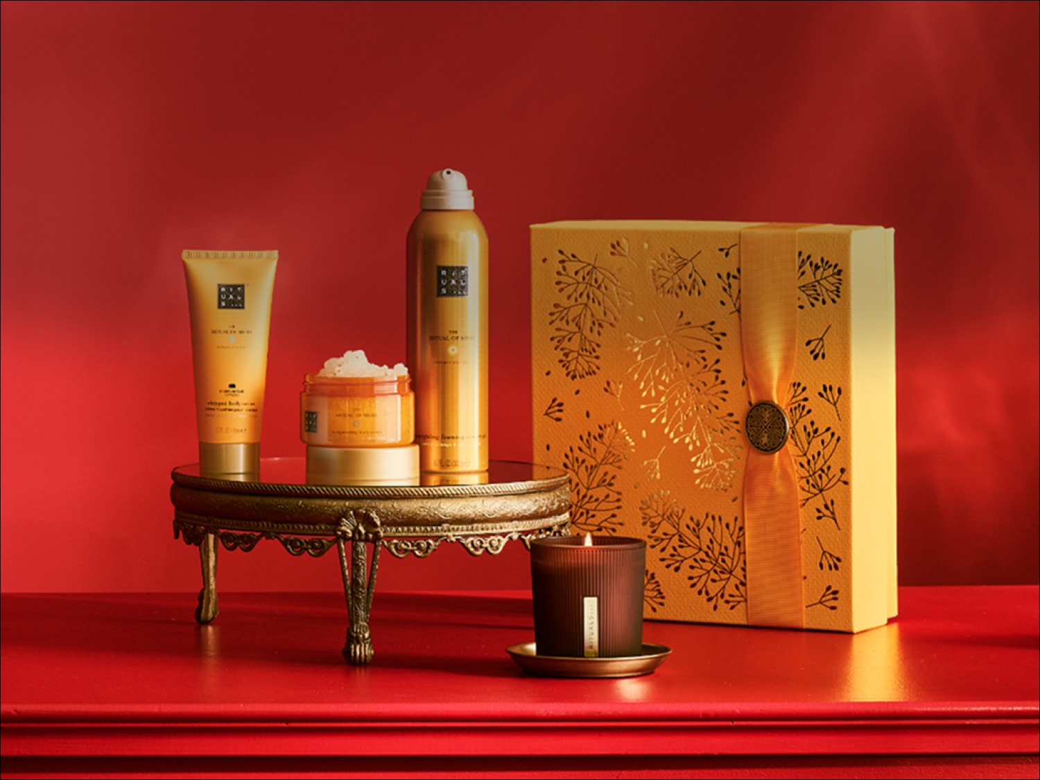Auksinės spalvos „Rituals of Mehr“ dovanų rinkinys su kūno kremu, putojančiu dušo geliu, kūno šveitikliu ir kvapia žvake ant auksinio staliuko raudoname fone.