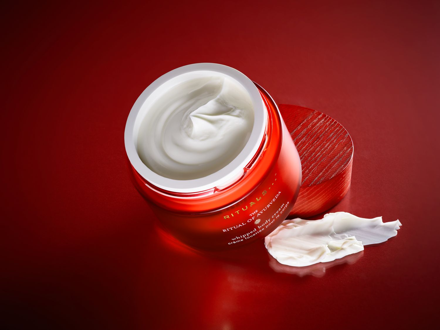 Odkryj krem do ciała Rituals of Ayurveda w czerwonym słoiczku z drewnianym wieczkiem, obok którego leży porcja kremu o gładkiej konsystencji.