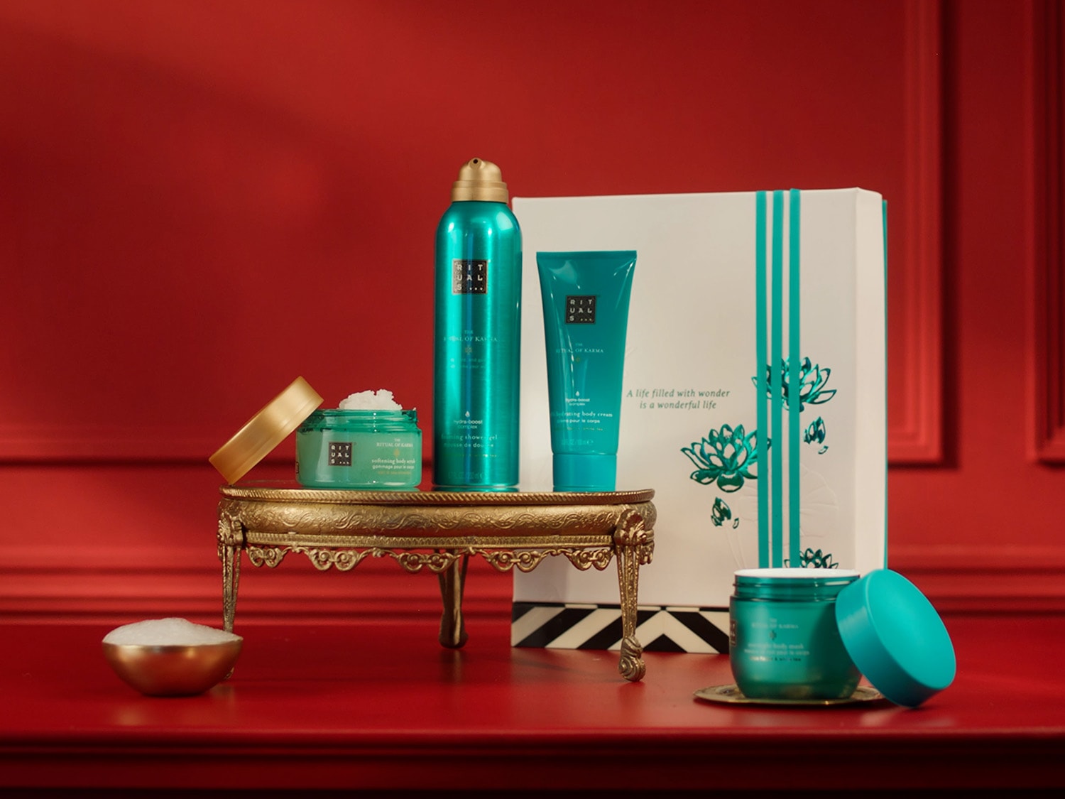 Prabangus „Rituals of Karma“ rinkinys su turkio spalvos pakuotėmis, įskaitant kūno kremą, dušo putas ir šveitiklį, ant auksinio staliuko, fone – baltas dovanų rinkinys su lotoso gėlėmis.