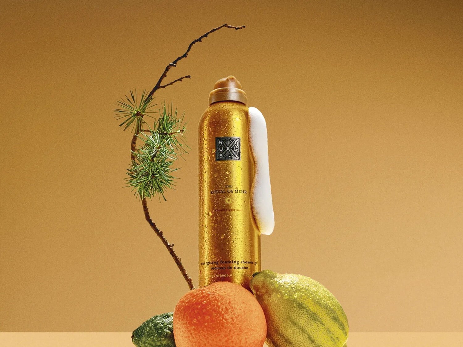 Złoty żel pod prysznic Ritual of Mehr z pianką, gałązką sosny i owocami cytrusowymi na złotym tle.