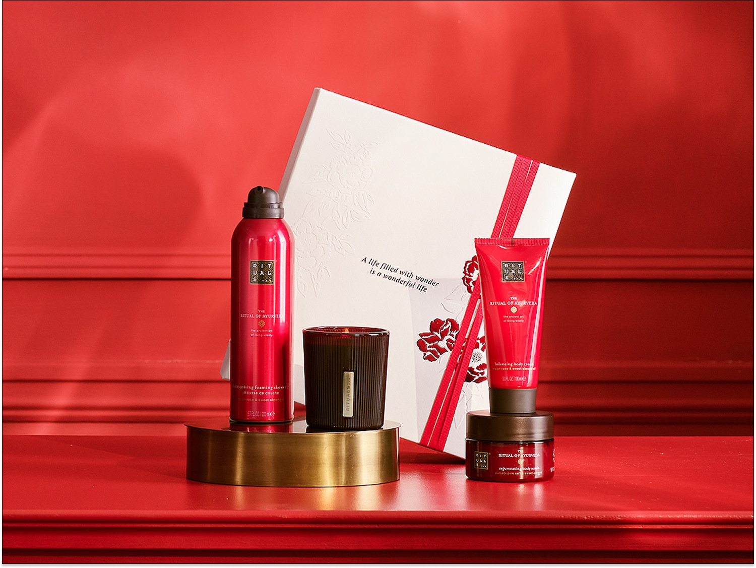 „Rituals of Ayurveda“ dovanų rinkinys su kūno kremu, dušo žele, kūno šveitikliu ir kvapniąja žvake, supakuotas į baltą dėžutę su raudonu kaspinu.