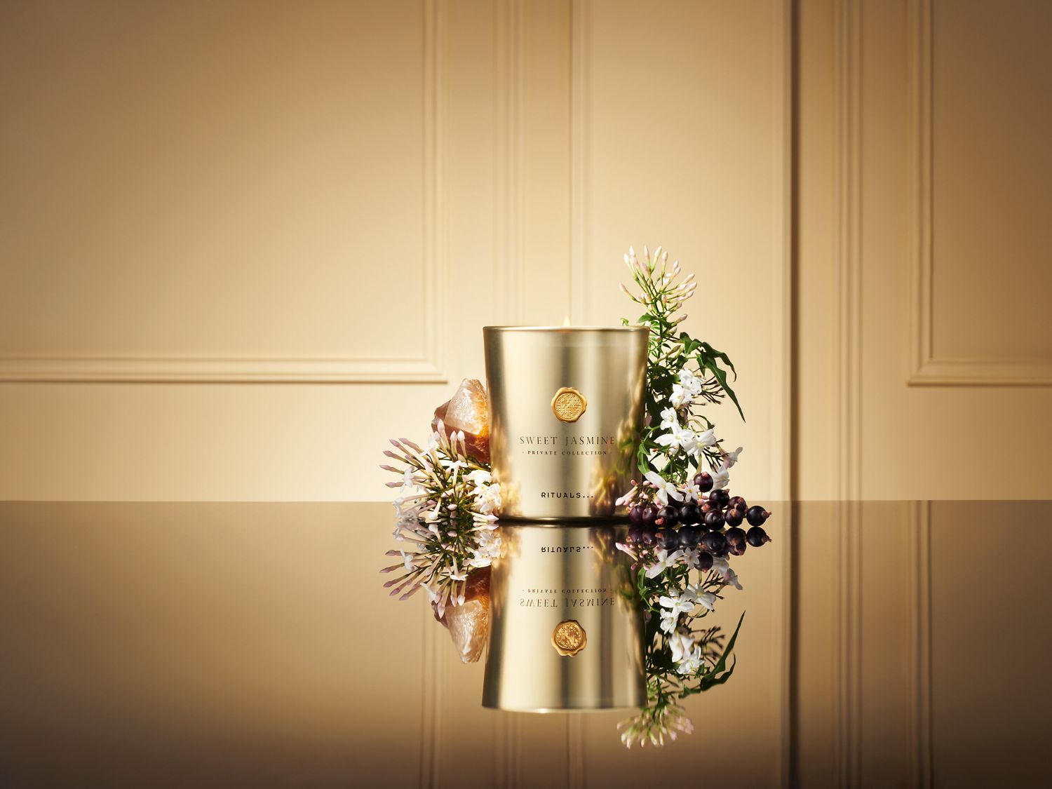 Świeca zapachowa Rituals Sweet Jasmine Private Collection w złotym opakowaniu, ozdobiona białymi kwiatami jaśminu i kamieniami, odbija się w błyszczącej powierzchni.