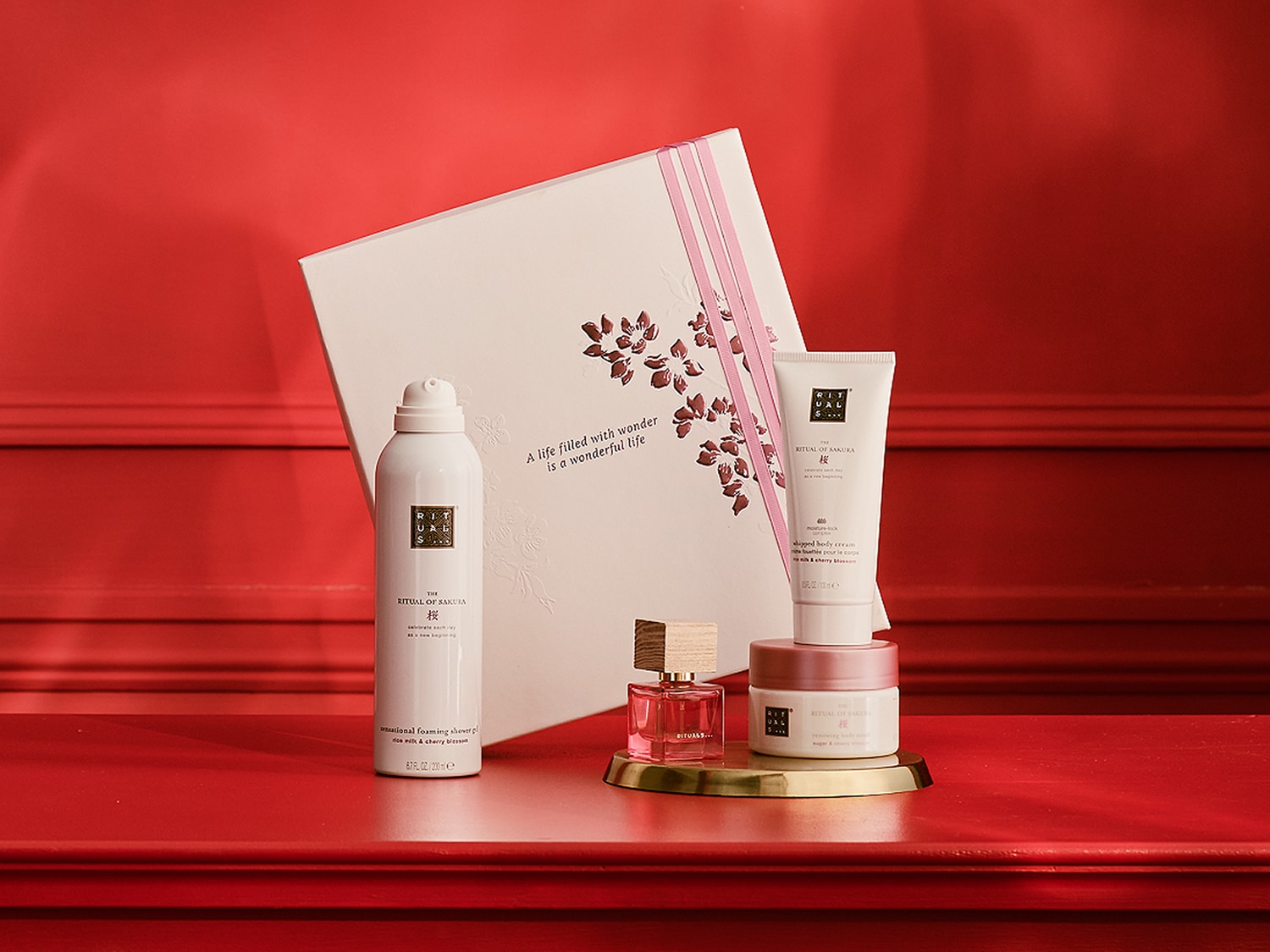 Šiame paveikslėlyje matome „Rituals of Sakura“ dovanų rinkinį, kuriame yra kūno kremas, dušo želė, kūno purškiklis ir kvepalai, visa tai ant raudono paviršiaus su atitinkamu fonu.