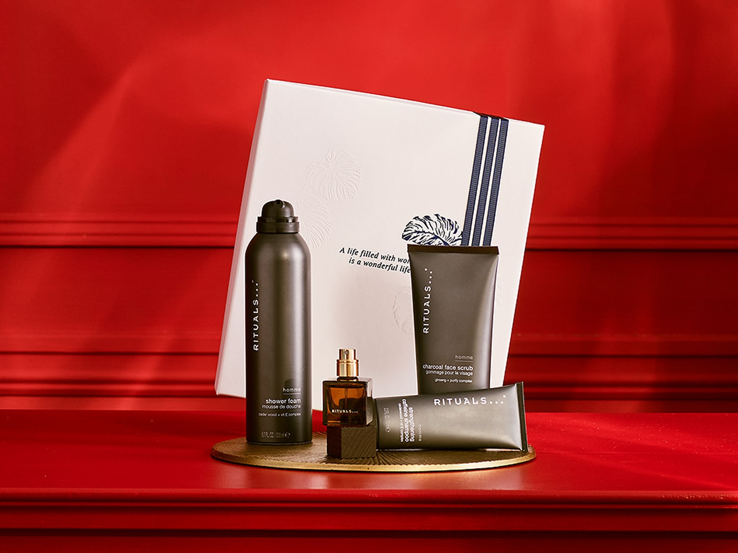 „Rituals... Homme“ rinkinys su juodu korpusu dušo putomis, veido šveitikliu, šampūnu ir odekolonu ant auksinės lėkštės, o fone – balta dovanų dėžutė su užrašu „Gyvenimas, kupinas darbo, yra nuostabus gyvenimas“.